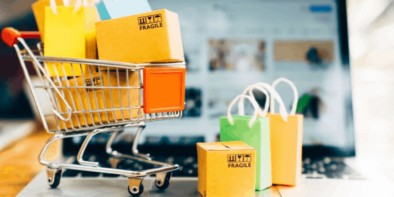 Notebook aberto com site de compras ao fundo e carrinho de cheio de sacolas e pacotes a frente.