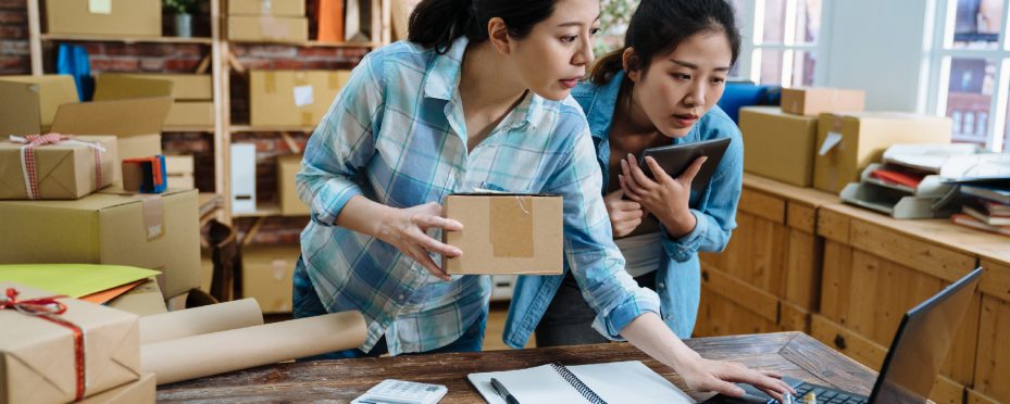 Duas mulheres em um estoque rodeadas por caixas olham um marketplace B2B em notebook sobre a mesa.
