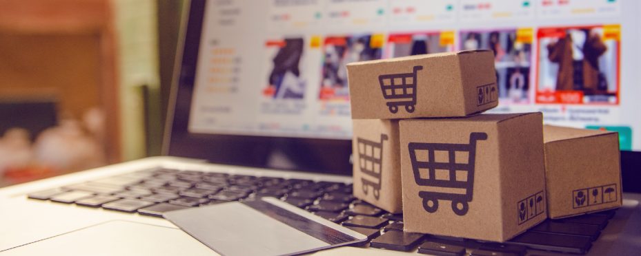Notebook aberto em site para anunciar produtos. Caixas de papelão com desenho de carrinhos sobre o teclado e a frente um cartão de crédito.