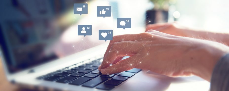 Notebook aberto com mãos digitando no teclado para anunciar na internet e ícones de redes sociais à frente.