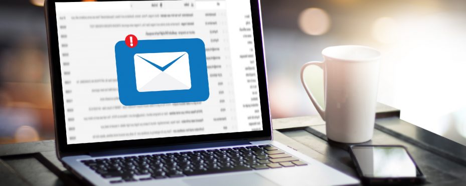 Notebook aberto em página de e-mails com ícone de envio. Ao Lado uma xícara branca e um celular preto.