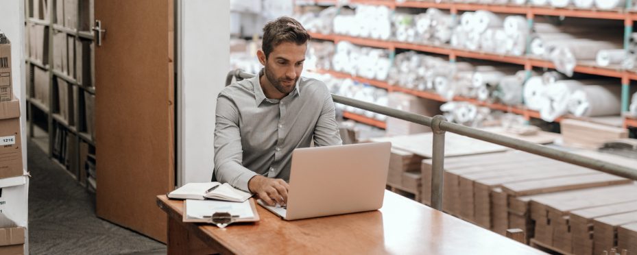 Homem em um estoque sentado em frente a notebook pesquisando sobre marketing industrial.