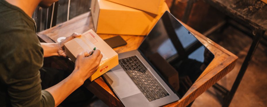 Homem sentado empacota produto vendido pelo portal B2B em uma mesa com notebook aberto.