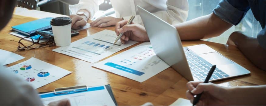 Pessoas fazendo anotações em folhas e notebook sobre cadeia de suprimentos.