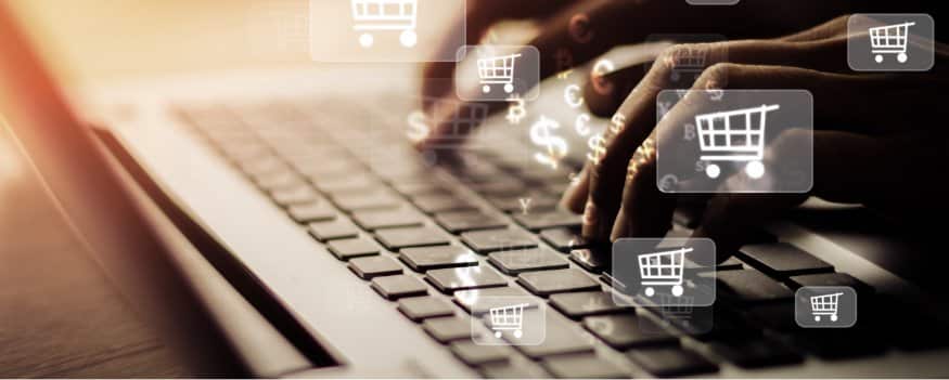 Mãos digitando em teclado de notebook e ícones de compras e cifrão em destaque.