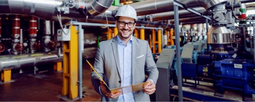 Homem com capacete branco em pé no meio das máquinas de uma indústria segurando pasta amarela com anotações sobre marketing para indústria.