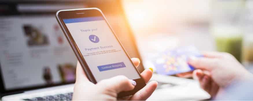 Mãos segurando celular em e finalizando o pagamento de produto feito em plataforma marketplace B2B. Ao fundo um notebook aberto e página do site.