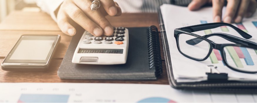 Mãos calculando balanço patrimonial de uma empresa em uma mesa com caderno, calculadora, celular, e óculos.