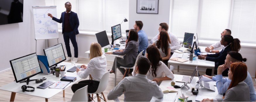 Grupo de pessoas reunidas em escritório e homem de pé apresentando treinamento sobre como lidar com a concorrência.