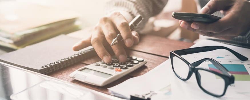 Homem usando a calculadora para fazer Cálculo markup em uma mesa com papéis, óculos e caderno.