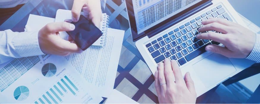 Mãos digitando em notebook e outras mexendo em celular procurando informações para conciliação bancária.