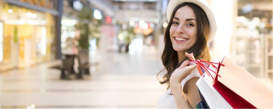 Mulher sorrindo com chapéu segurando sacolas após fazer compras em shopping.