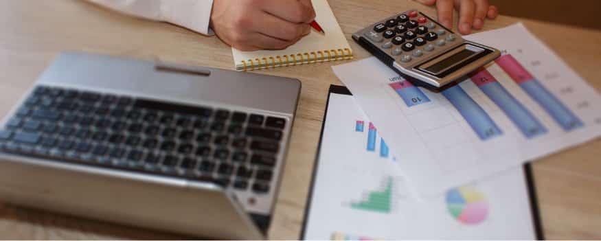 Uma pessoa fazendo anotações em um papel com a calculadora enquanto consulta o computador e gráficos.